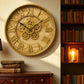 Decorative wall clock with gear design and Roman numerals in a room with books and a lamp.